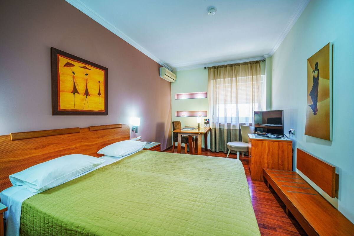 Ein Hotelzimmer mit einem großen Bett, einem Schreibtisch, einem Sessel, einem Fernseher und Kunstwerken an den Wänden.