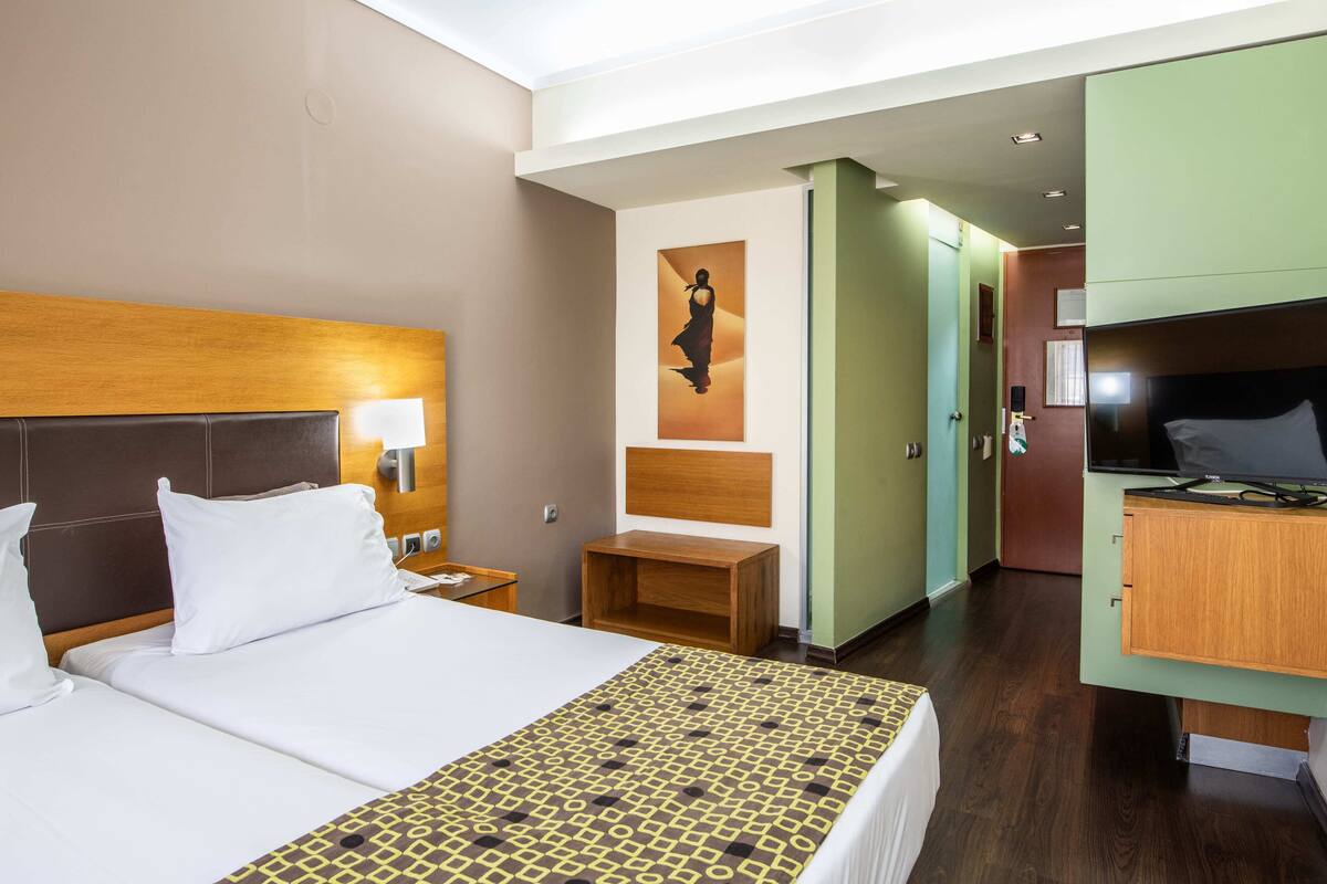 Ein Hotelzimmer mit einem großen Bett, einem Holz-Kopfteil, einem Nachttisch mit Lampe, einem kleinen Holzbeistelltisch, einem an der Wand befestigten Fernseher und einem grünen Kleiderschrank.