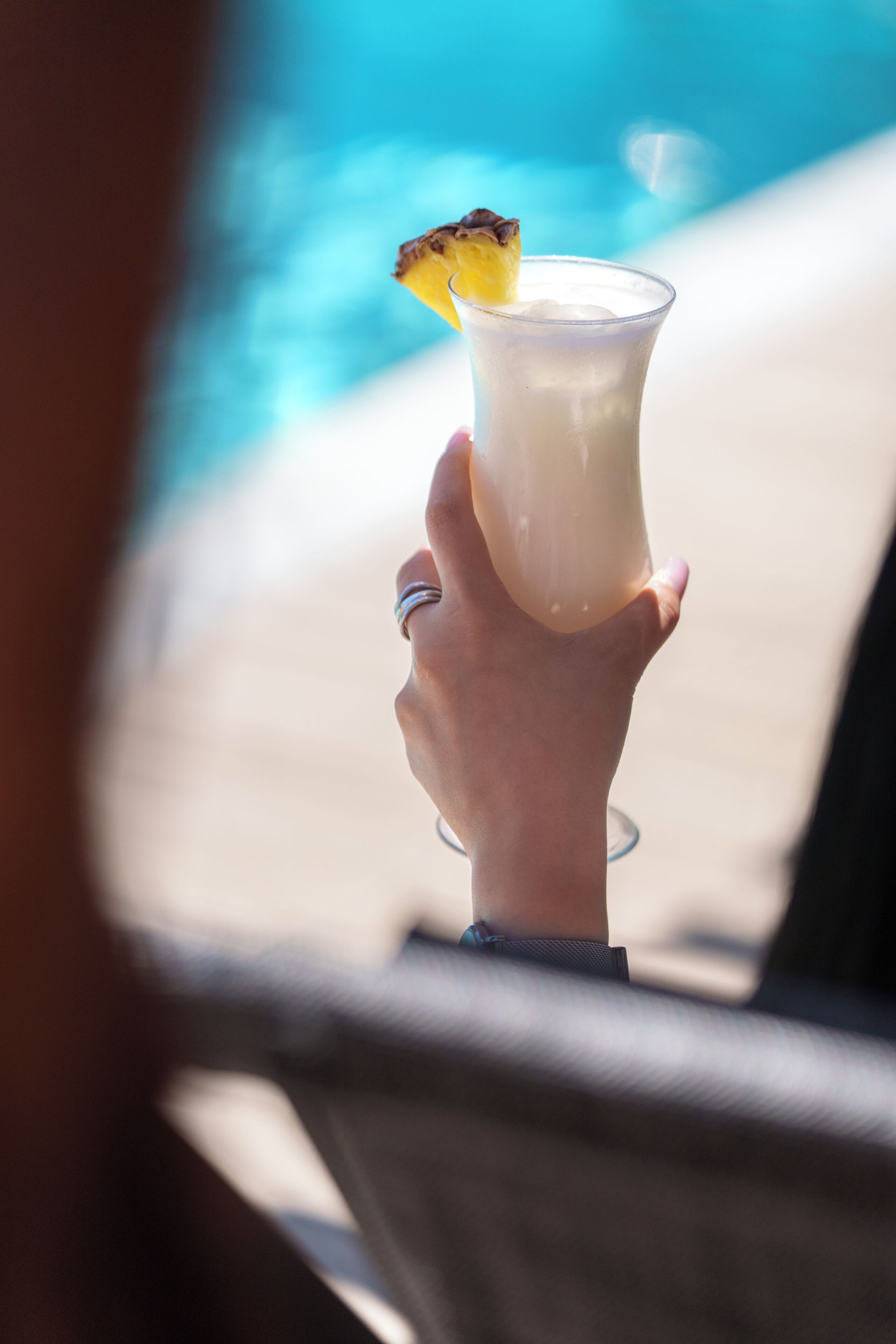 A hand holding a drink with a garnish, near a poolside.