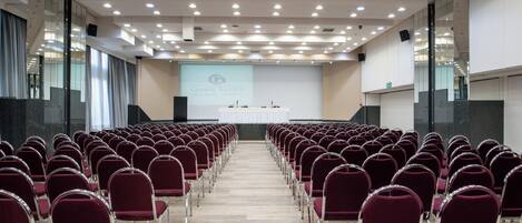 A conference room with rows of red chairs, a stage with a presentation screen, and a modern ceiling with recessed lighting.