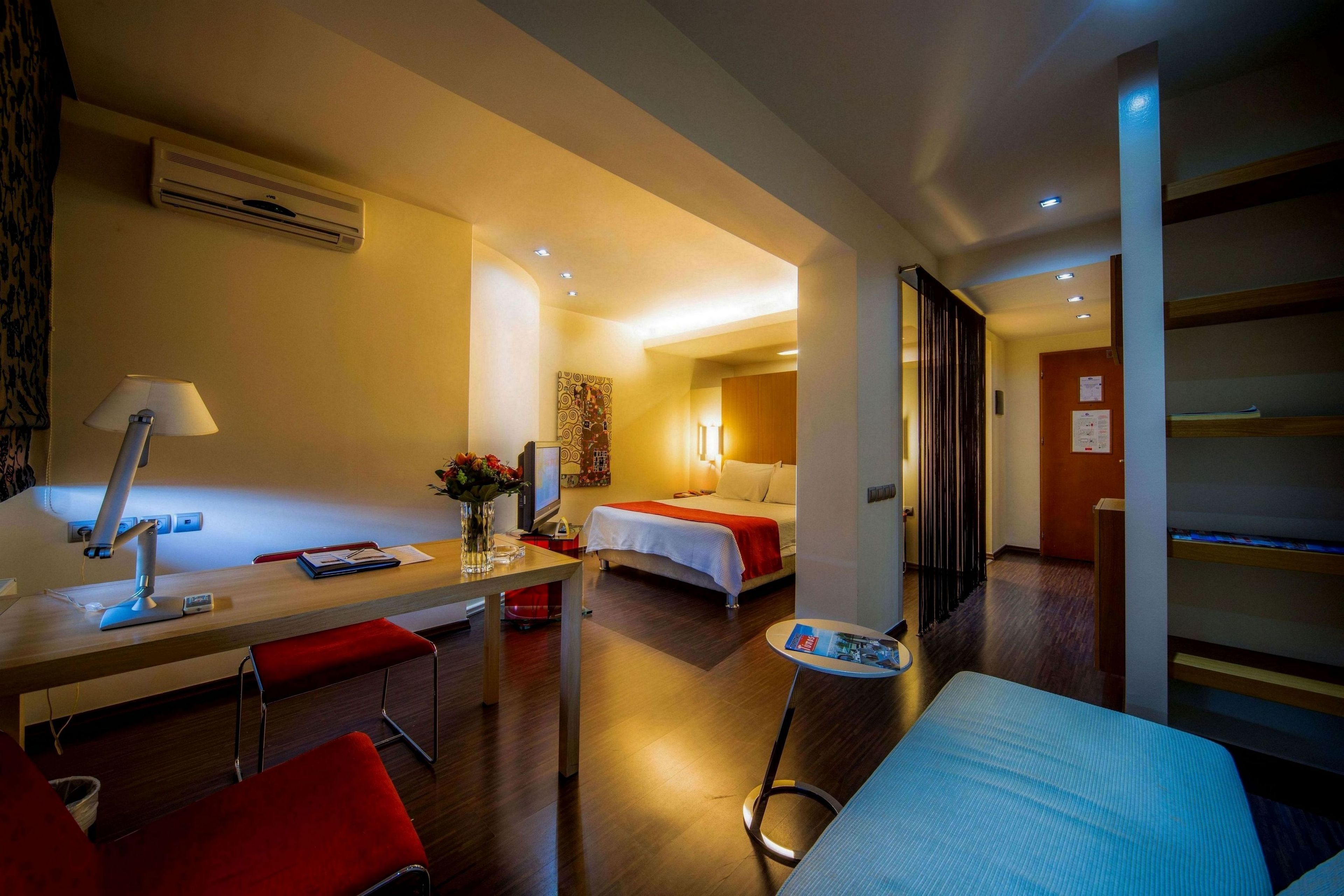 Ein Hotelzimmer mit Schreibtisch, zwei roten Stühlen, einem Bett, einem kleinen Tisch und einem Nachttisch mit Lampe.