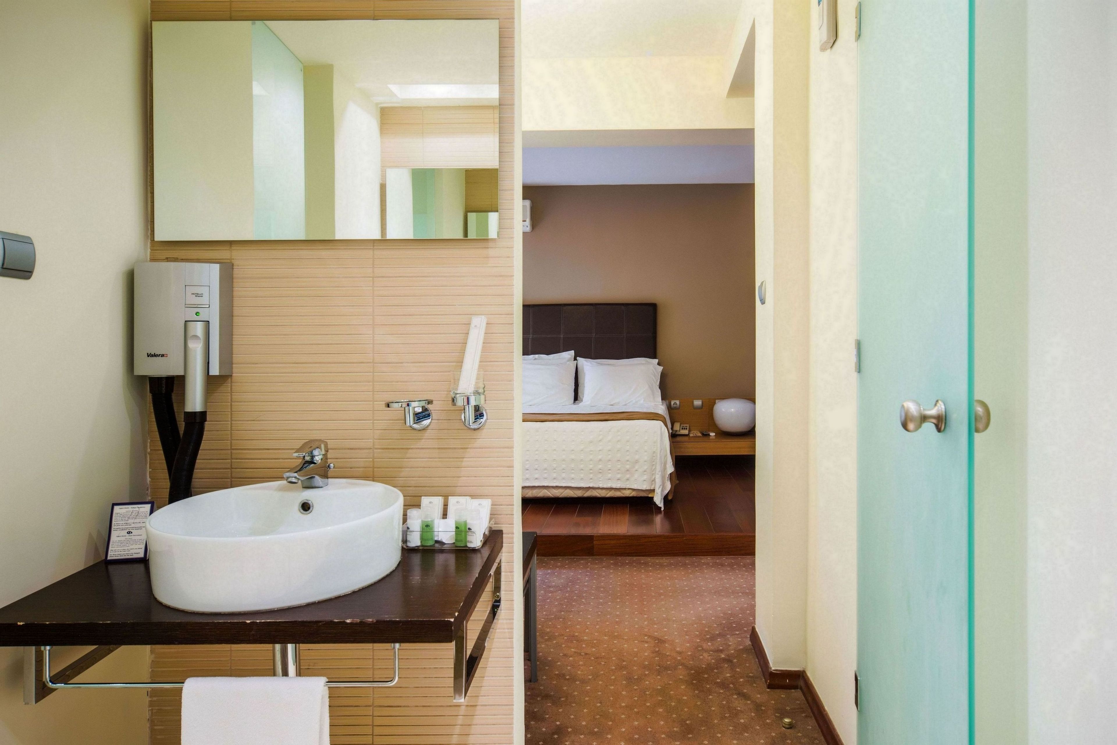 Une salle de bain d’hôtel avec un lavabo, un miroir et des articles de toilette. Une chambre à coucher avec un lit et une table de chevet est visible à travers une porte ouverte.
