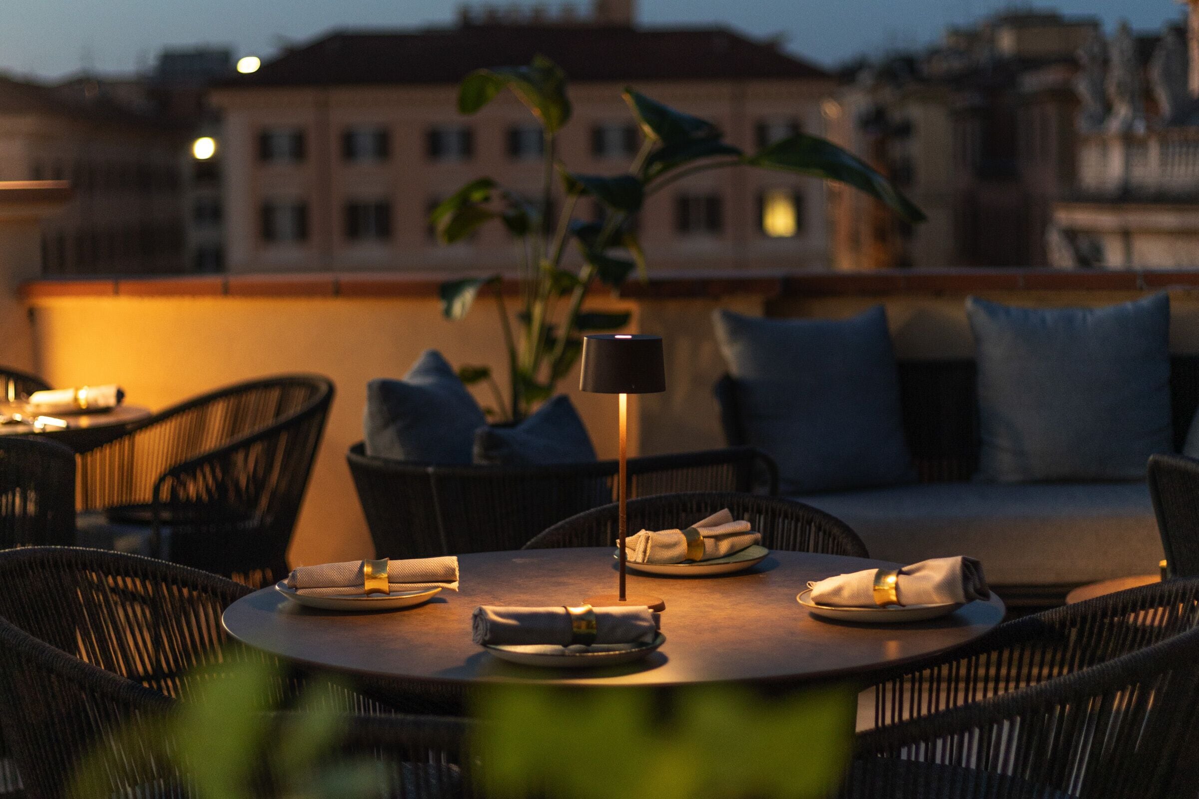 A rooftop terrace with round tables, wicker chairs, and a potted plant.