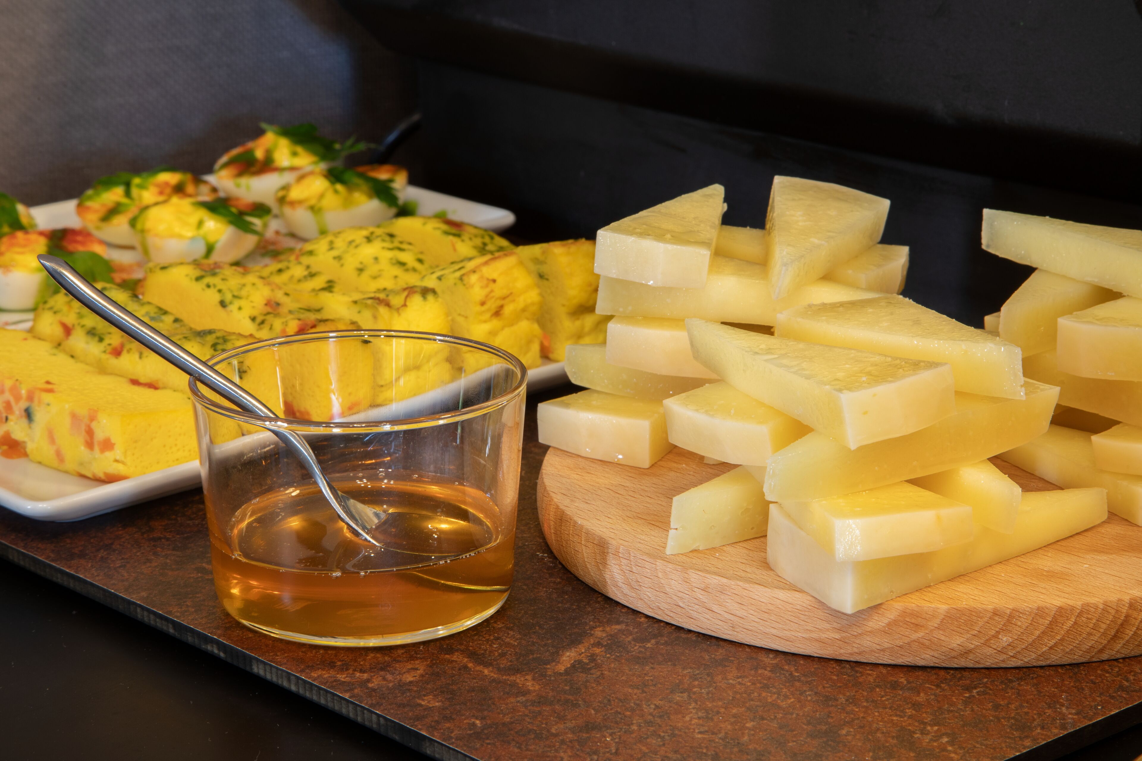 Une planche à découper en bois, avec des tranches de fromage et un verre de miel.