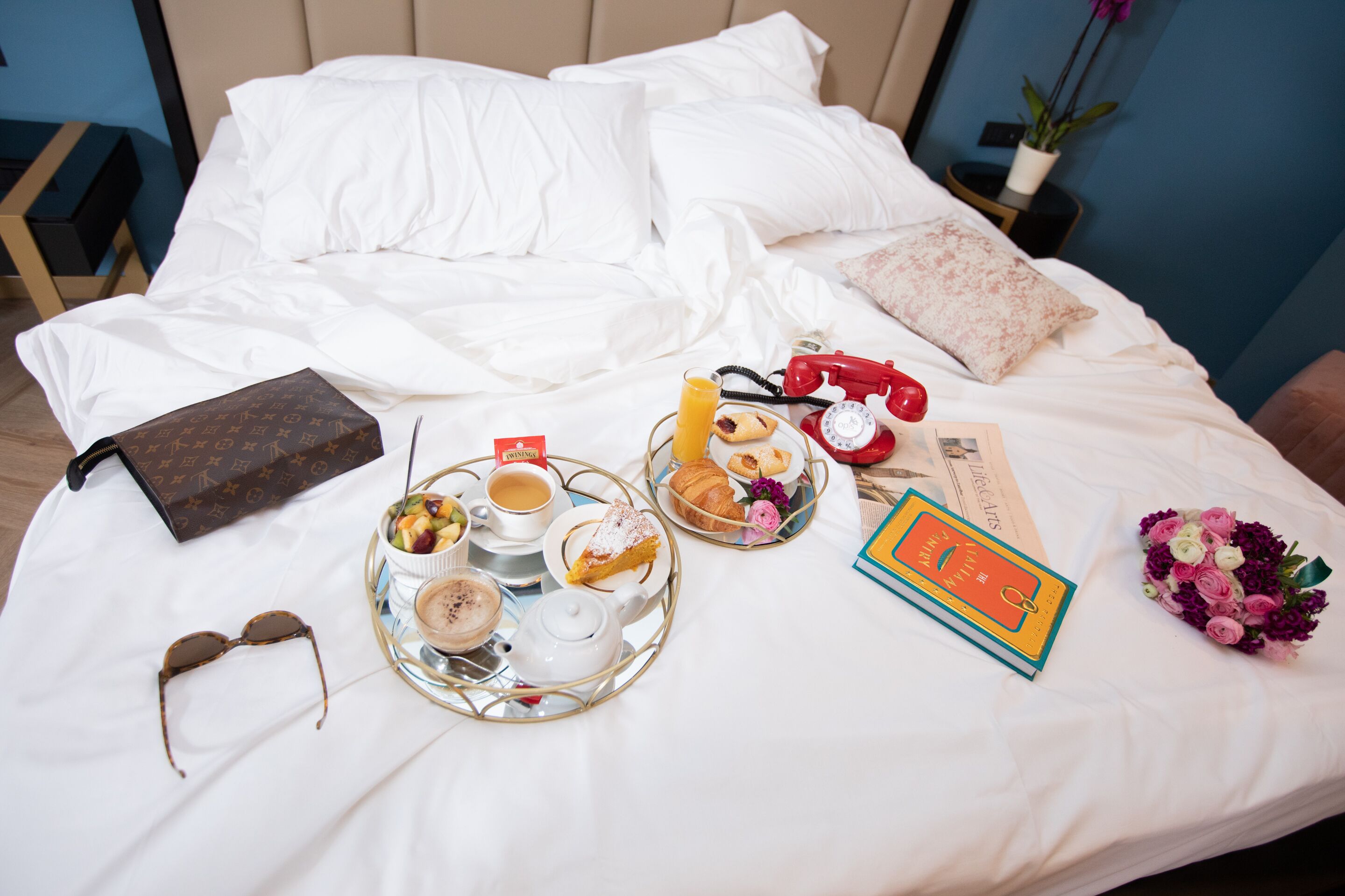 Una cama bien hecha con sábanas blancas, un bolso Louis Vuitton, una bandeja con alimentos para el desayuno, un ramo de flores y un libro.