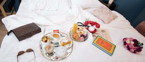 A neatly made bed with white linens, a Louis Vuitton bag, a tray with breakfast items, a bouquet of flowers, and a book.