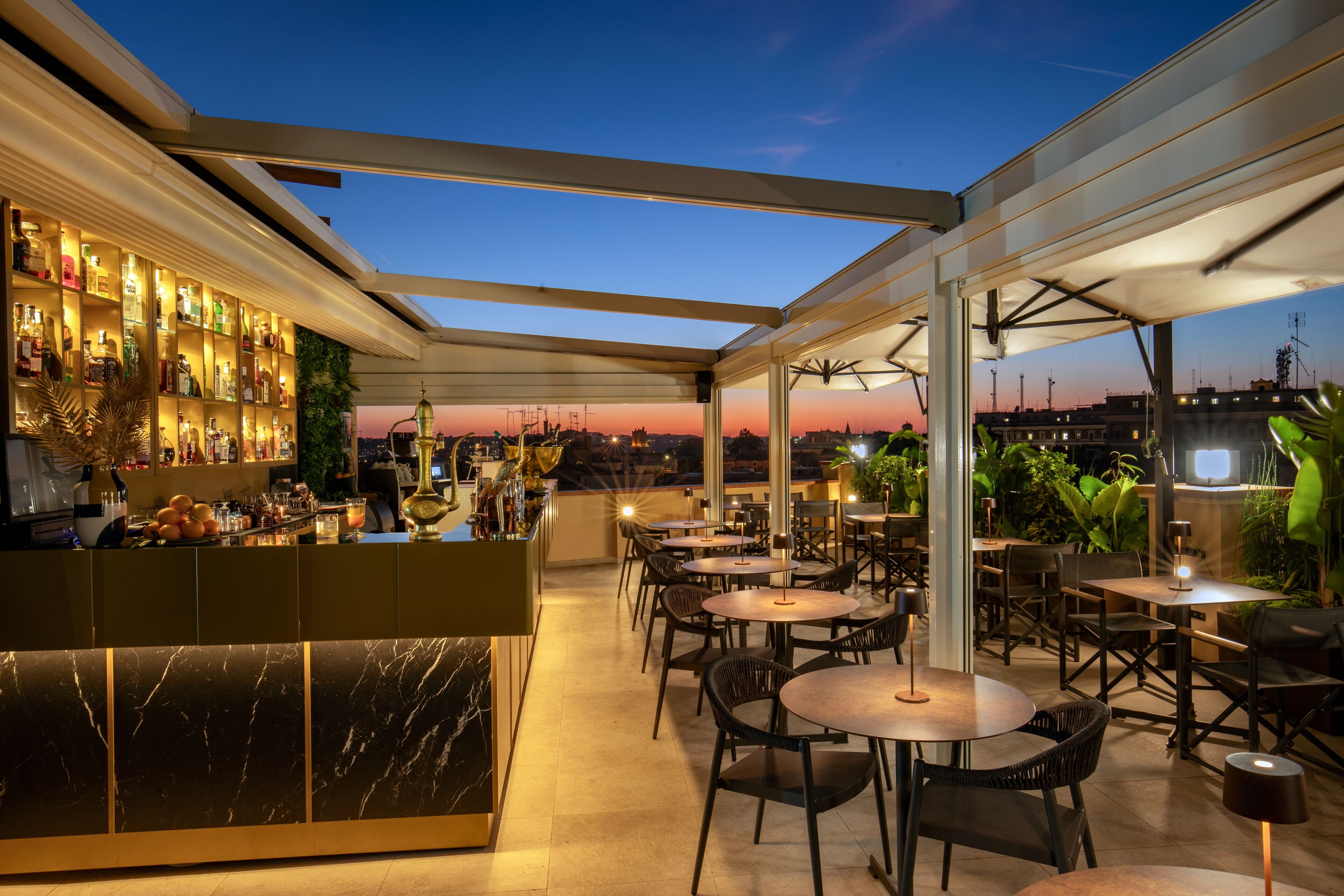 Un bar situé sur un toit, avec un comptoir, un espace salon et une vue sur la ville au crépuscule.