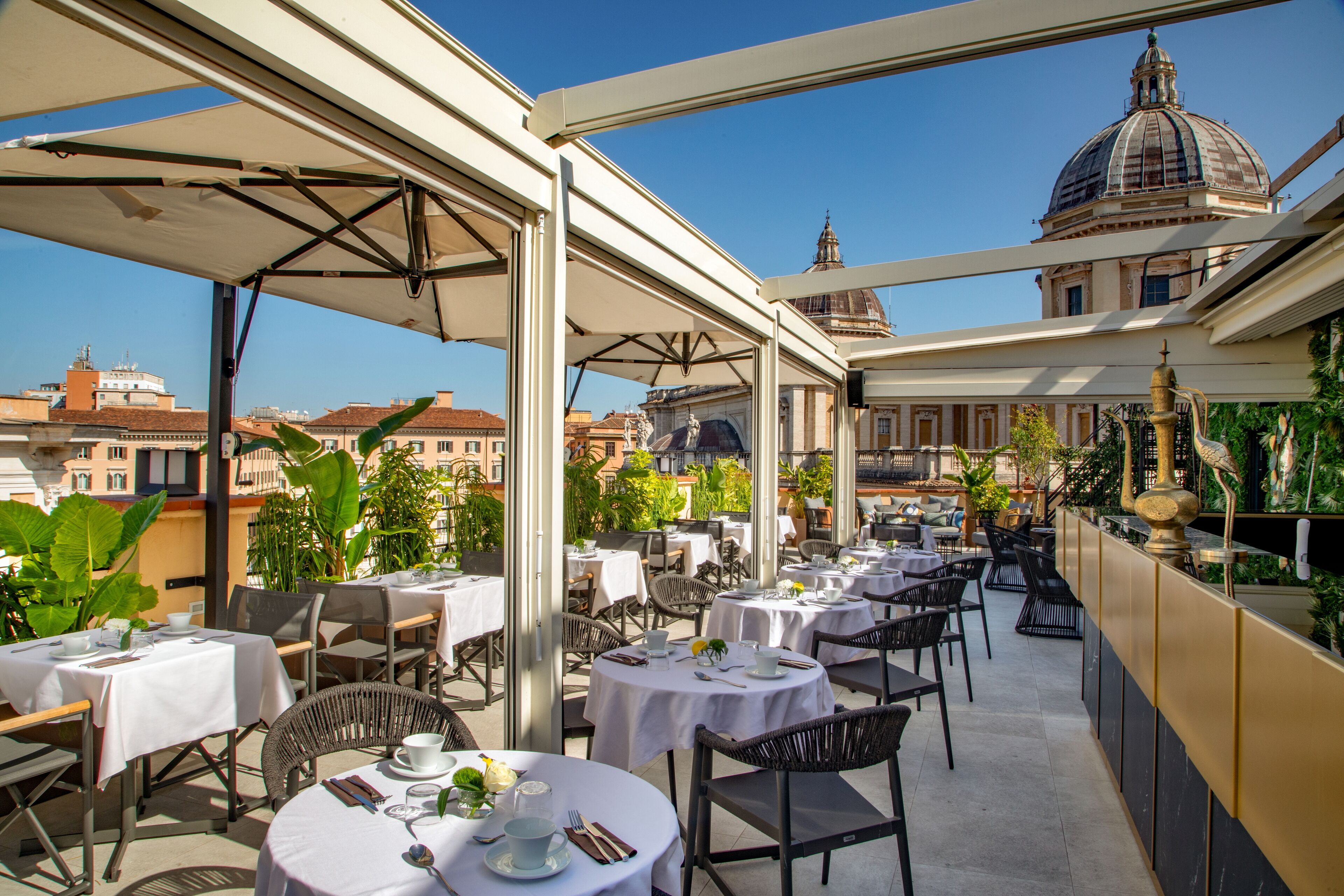 Zona pranzo all'aperto con tavoli e sedie rotonde, una sezione coperta e vista su un edificio storico con una cupola.