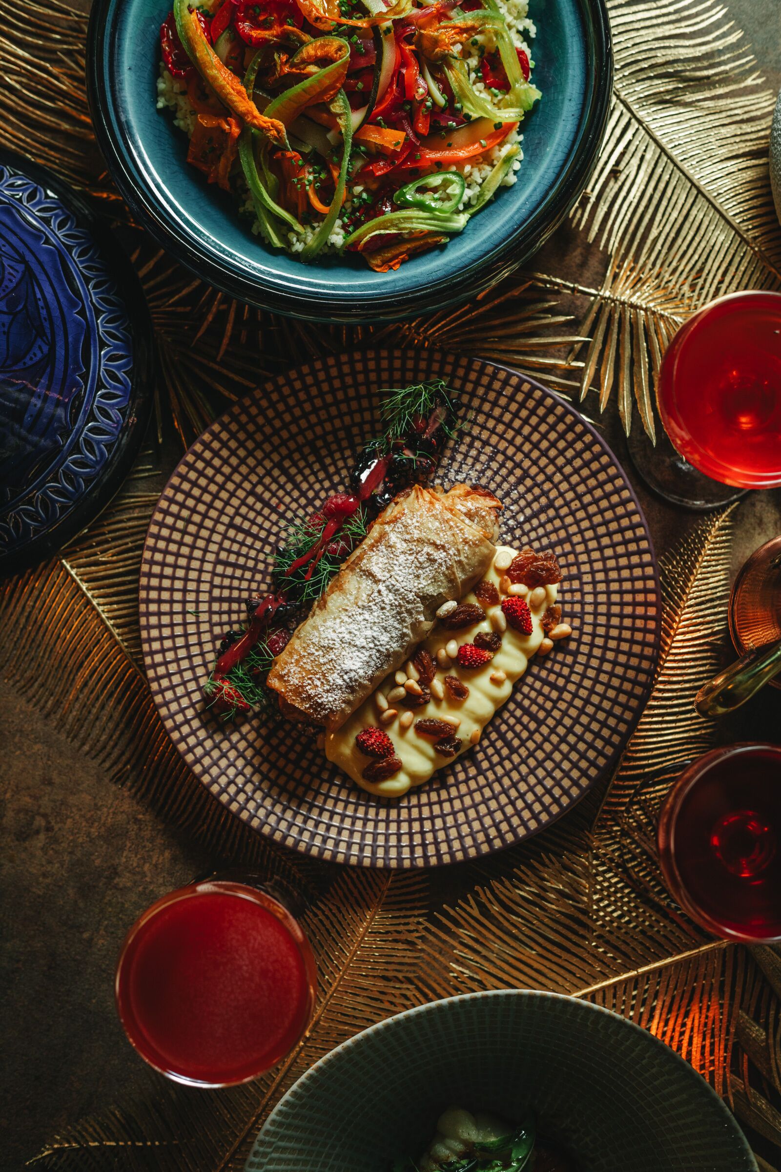 A table with a blue plate of food, a patterned plate with a dessert, and two glasses of red drink.