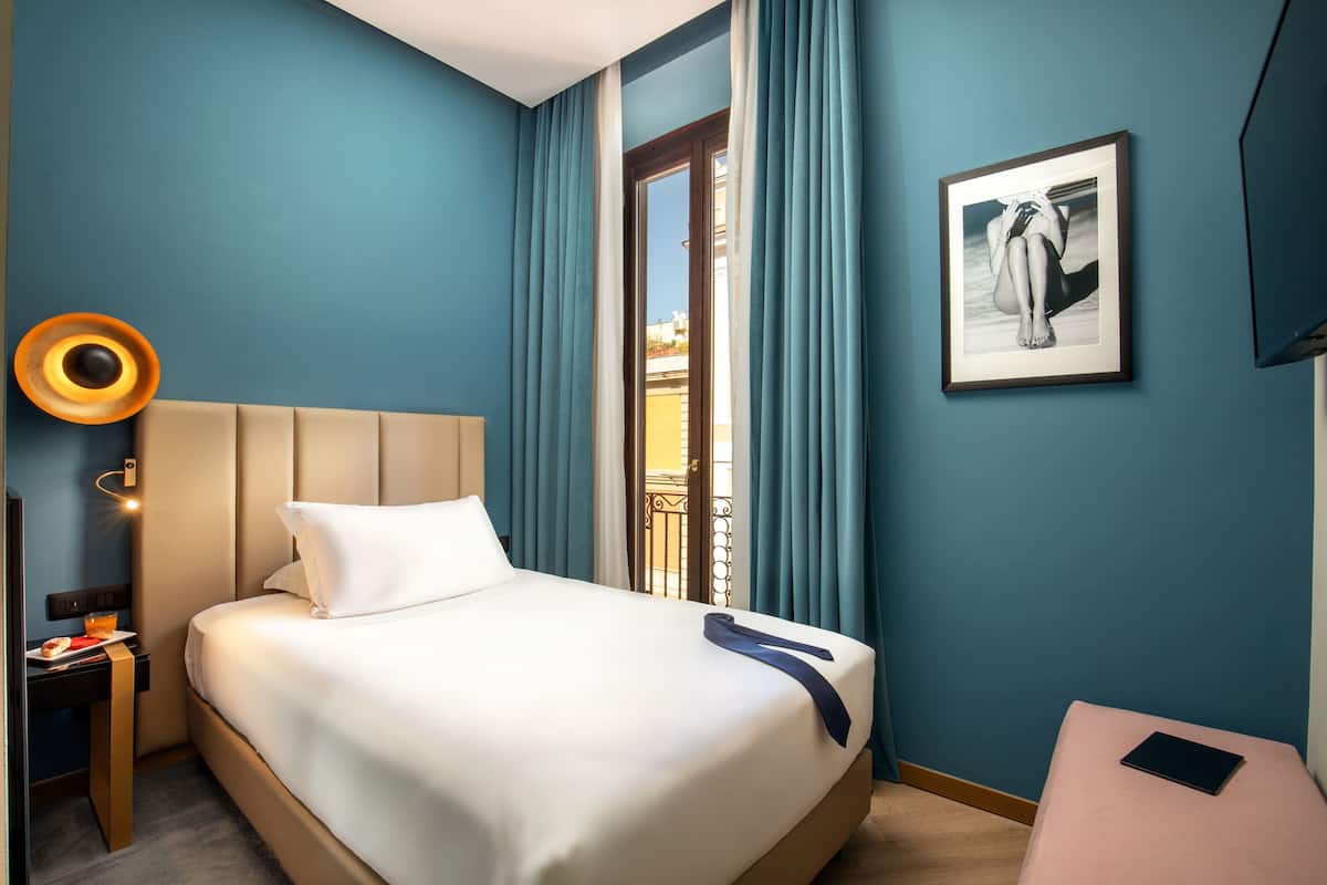 Une chambre d’hôtel avec un lit, une table de chevet sur laquelle se trouve une assiette de nourriture, un tableau encadré au mur et une fenêtre avec des rideaux.