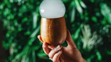 Uma mão segurando uma caneca de madeira com espuma, contra um fundo desfocado de folhas verdes.