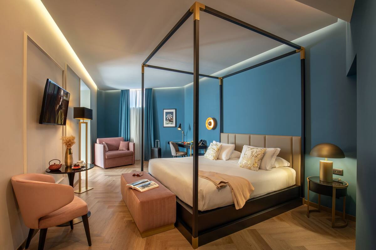 Une chambre moderne avec un lit à baldaquin, un coin salon comprenant un fauteuil et un pouf, un bureau avec une chaise et un téléviseur fixé au mur.