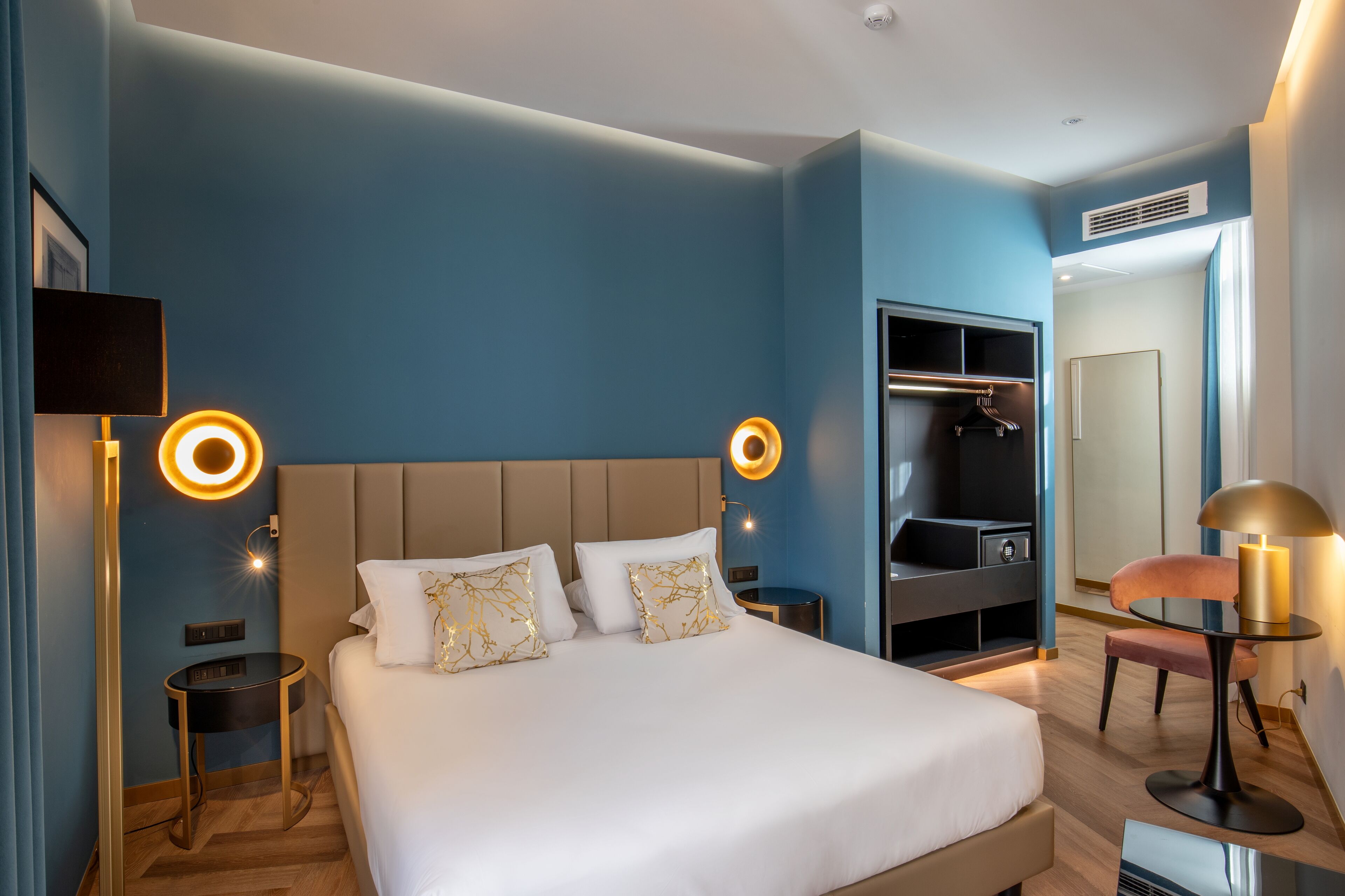 A modern hotel room with a large bed, bedside tables, a wardrobe, and a mirror.