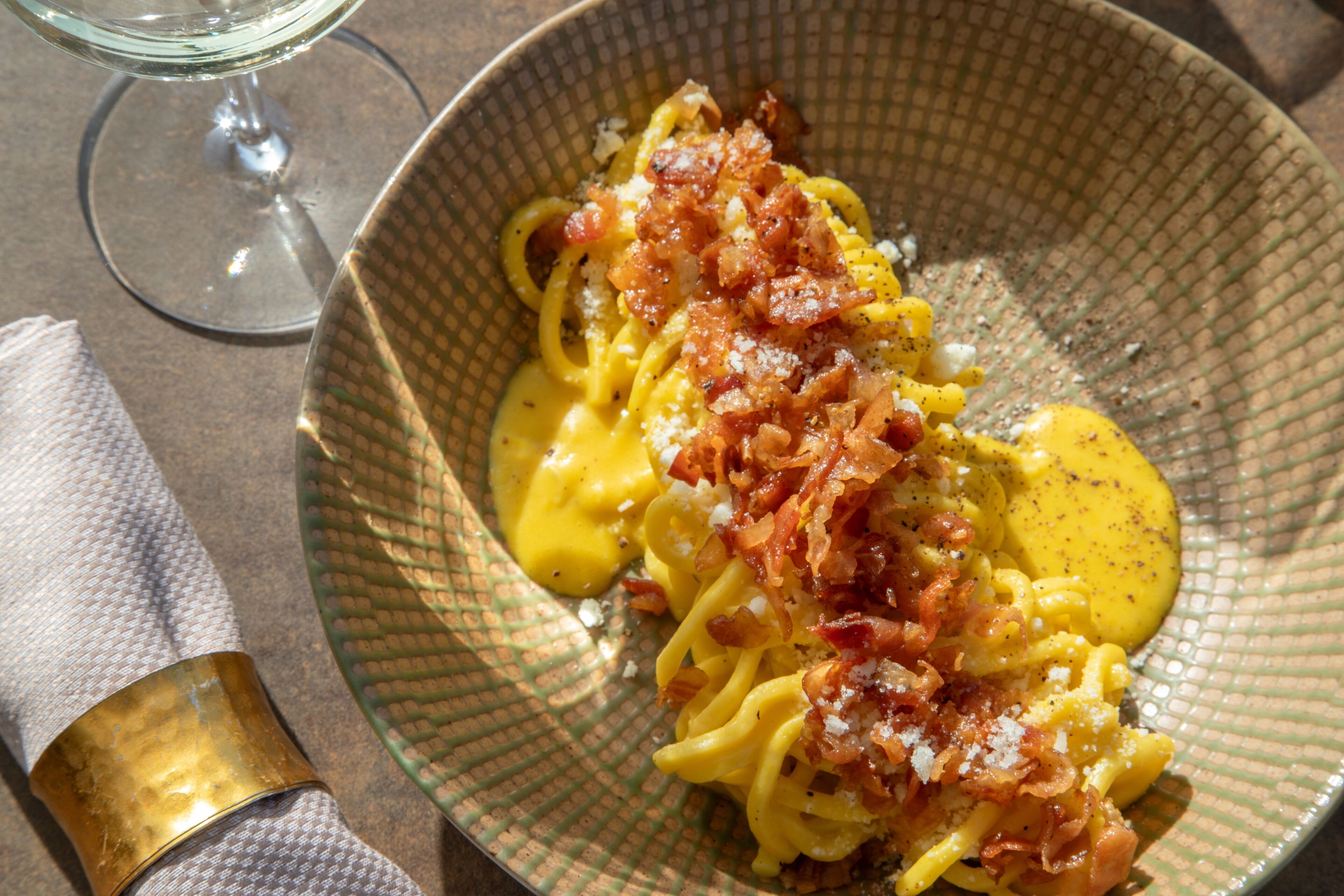 Una ciotola di pasta con bacon e formaggio, un bicchiere di vino e un tovagliolo su un tavolo.