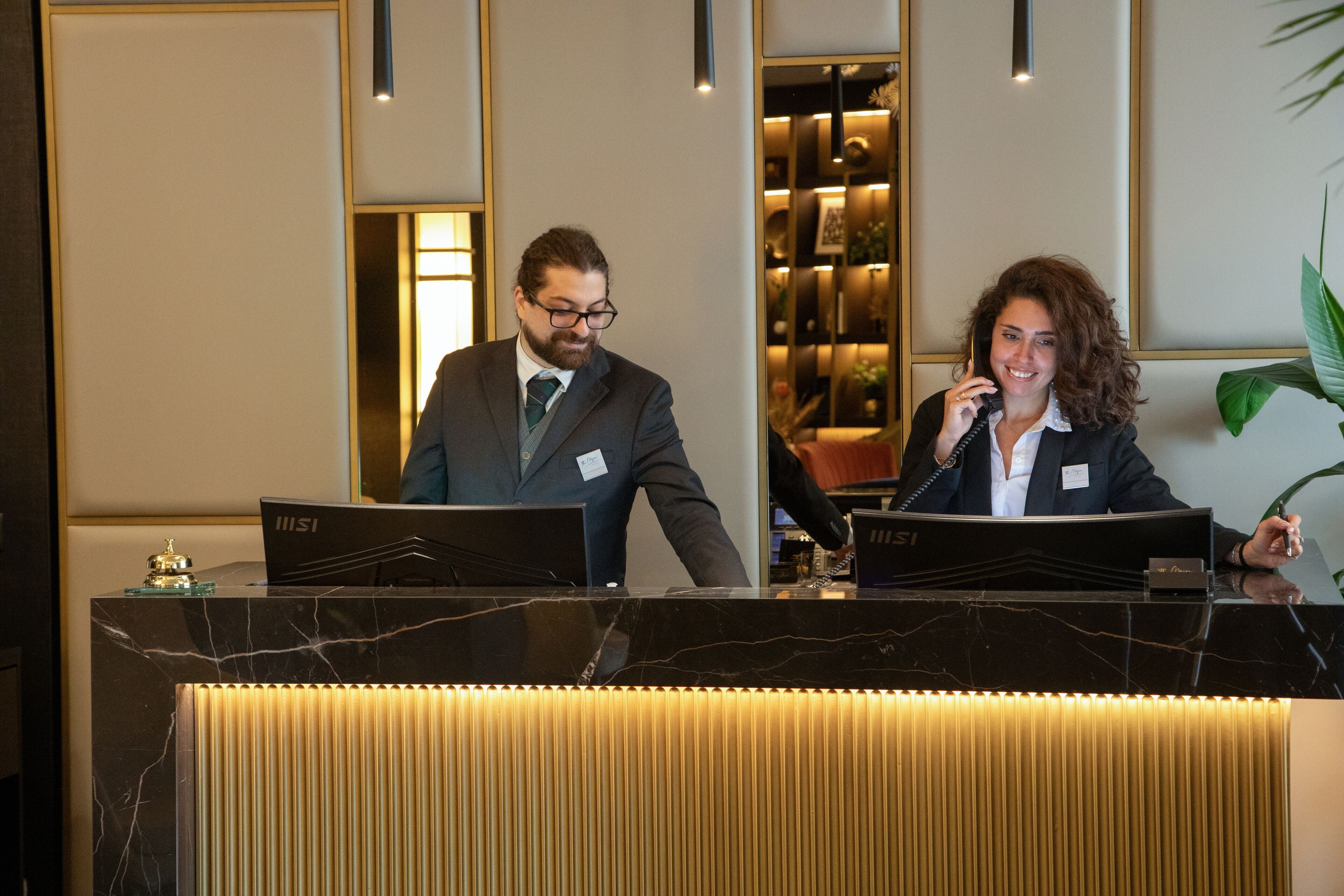 Una reception con due addetti, un bancone in marmo e un arredamento moderno con una pianta e quadri appesi.