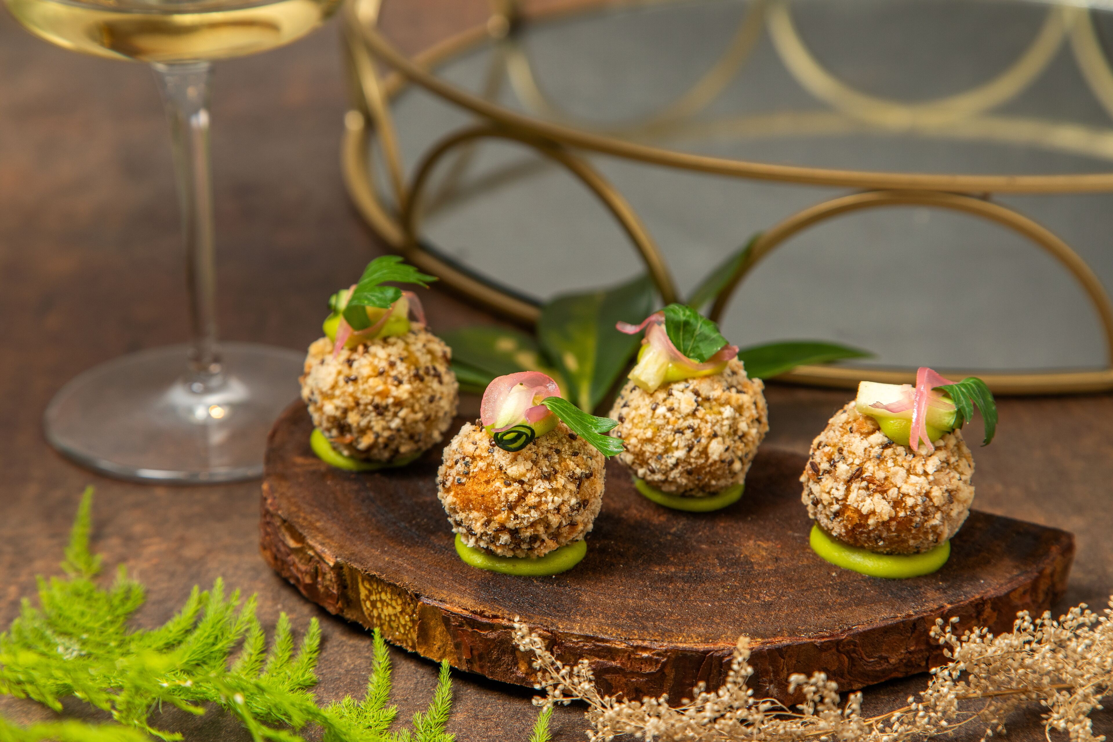 Four food items on a wooden board with a glass of white wine in the background.