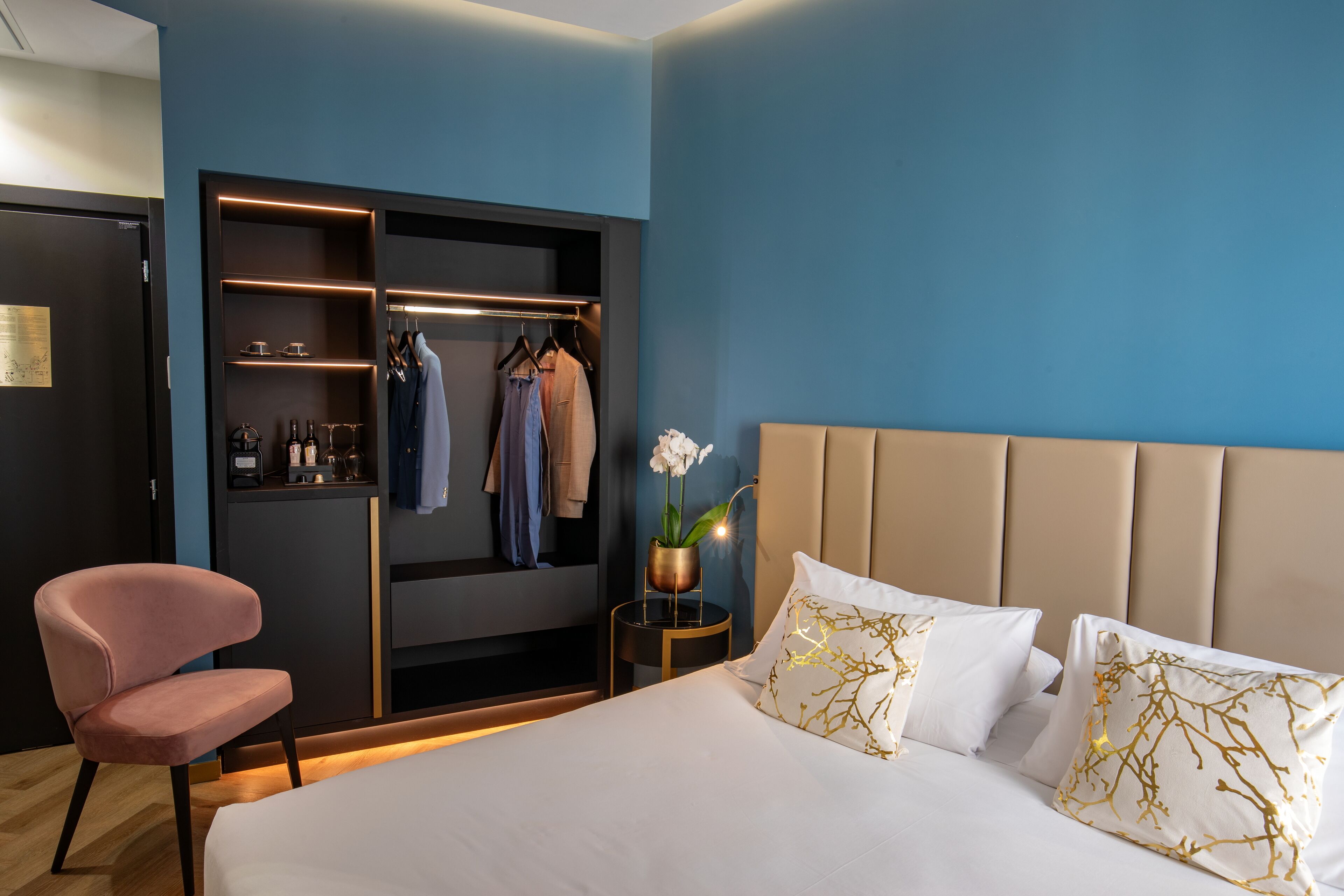 Un dormitorio con cama, una silla, armario y mesita de noche con lámpara y planta.