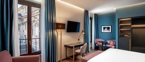 A hotel room with a bed, a desk with a chair, a TV, and a view of a building through a window.