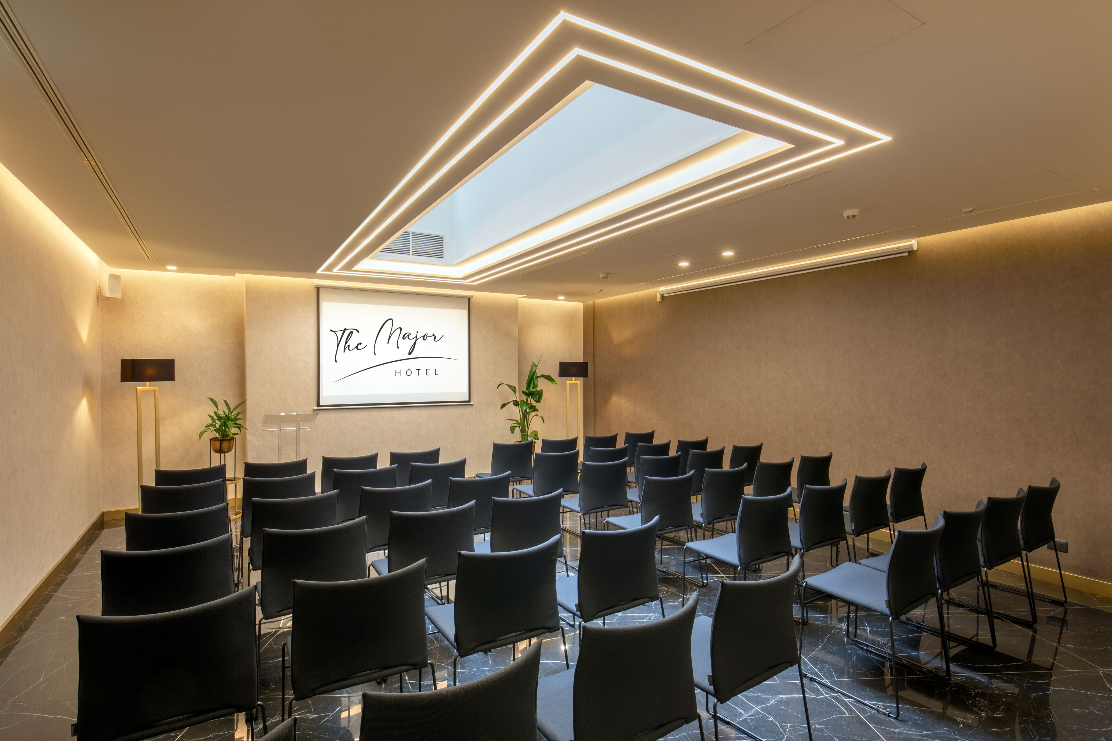 A conference room with rows of black chairs, a large screen displaying "The Major Hotel", and a skylight ceiling.