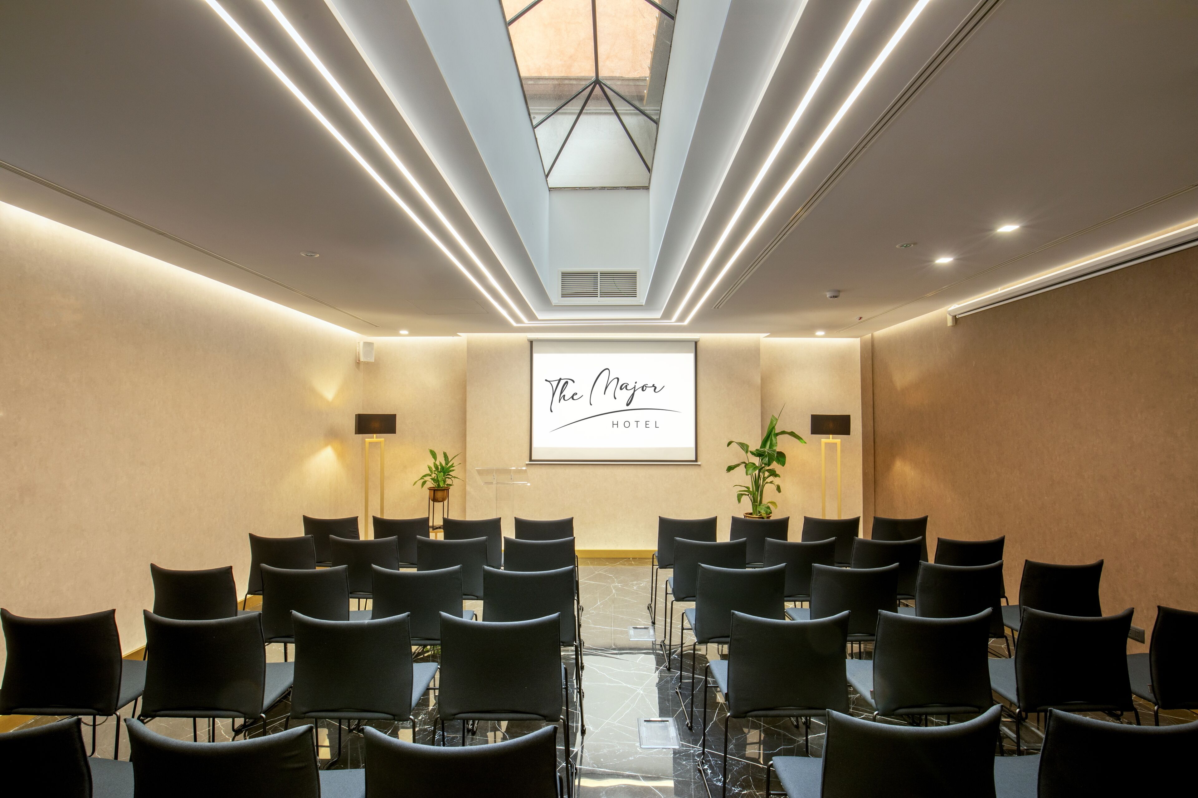 A conference room with rows of black chairs, a large screen displaying "The Major Hotel", and a skylight above.