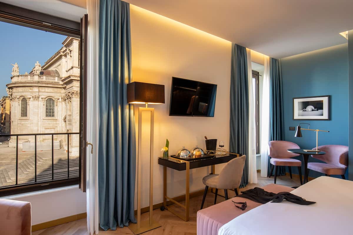 Une chambre d’hôtel avec une grande fenêtre, un bureau avec une lampe, deux fauteuils roses, un lit et un tableau encadré au mur.