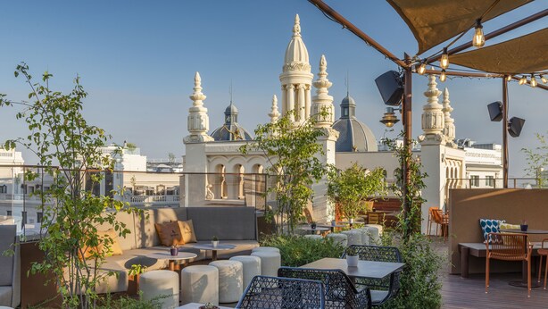 Eine Dachterrasse mit Sitzgelegenheiten im Freien, Blick auf ein weißes Gebäude mit Kuppeln und Lichterketten.