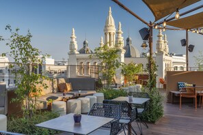 Eine Dachterrasse mit Sitzgelegenheiten im Freien, Blick auf ein weißes Gebäude mit Kuppeln und Lichterketten.