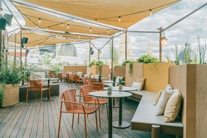 An outdoor patio with wooden flooring, round tables, and orange chairs. There are potted plants and a seating area with cushions.