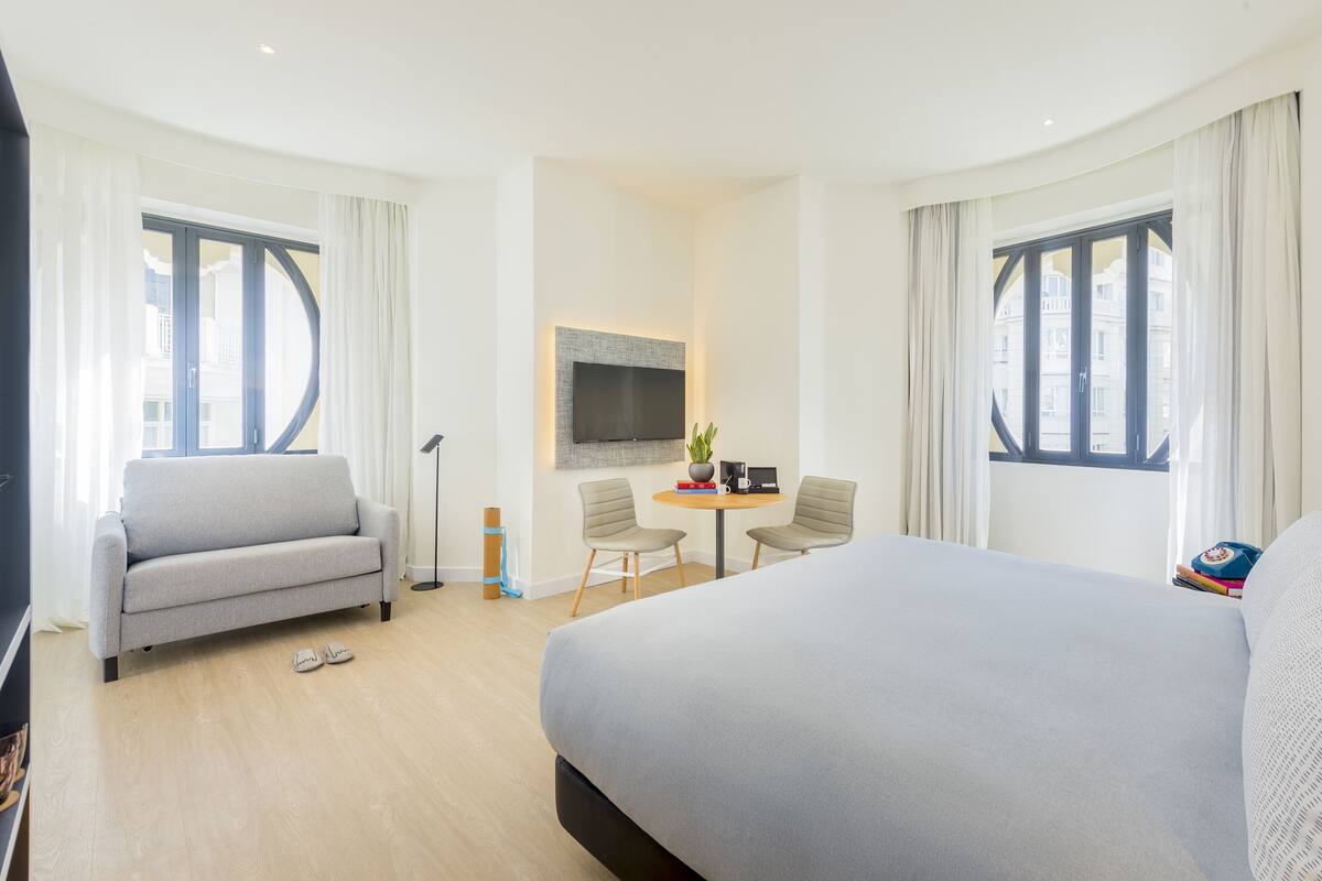 Camera d'albergo con un letto grande, un divano, un tavolino con sedie, una TV a schermo piatto montata a parete e ampie finestre con tende.