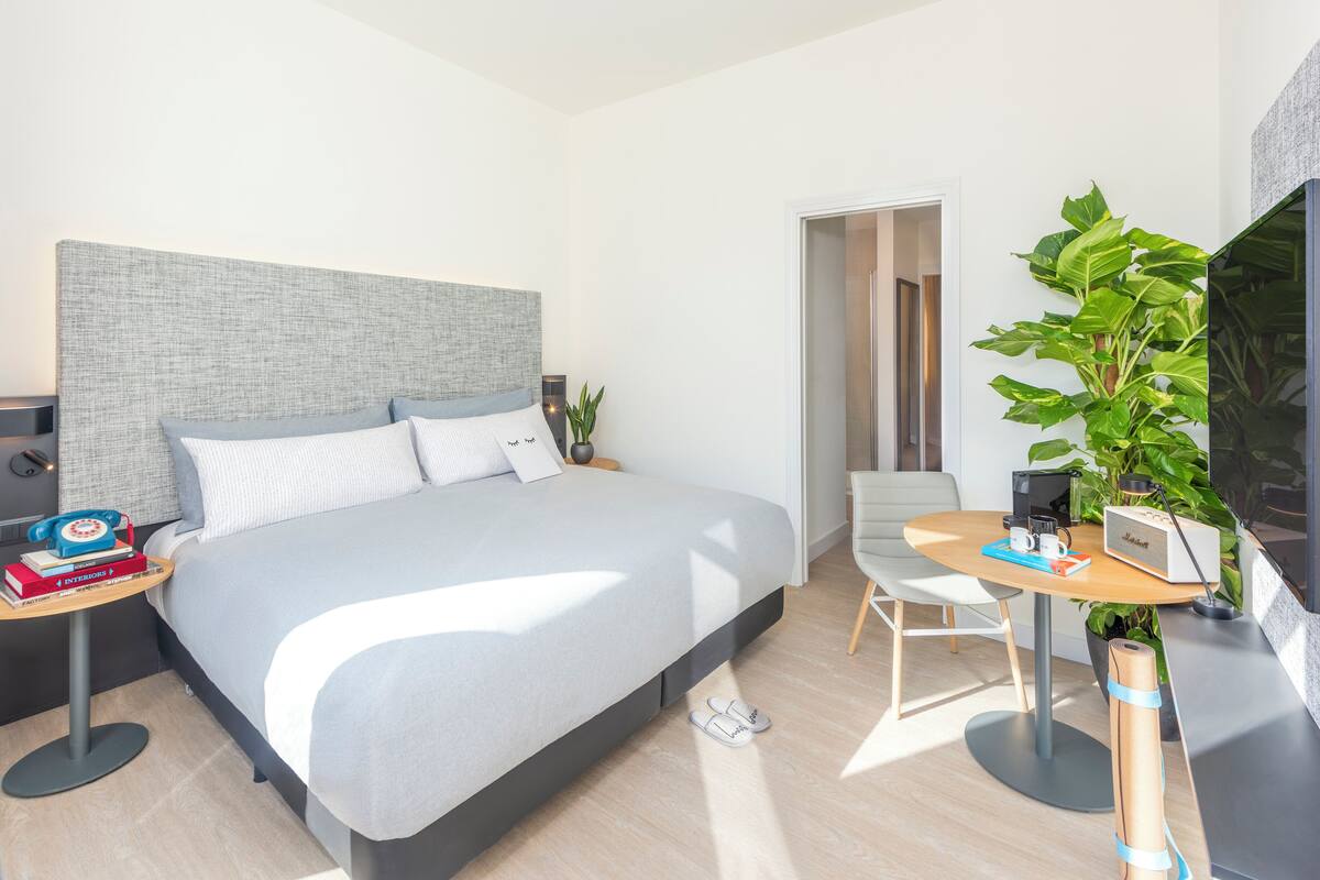 Ein modernes Hotelzimmer mit einem großen Bett, Nachttischen, einem Schreibtisch mit Stuhl, einem Fernseher und einer Zimmerpflanze.