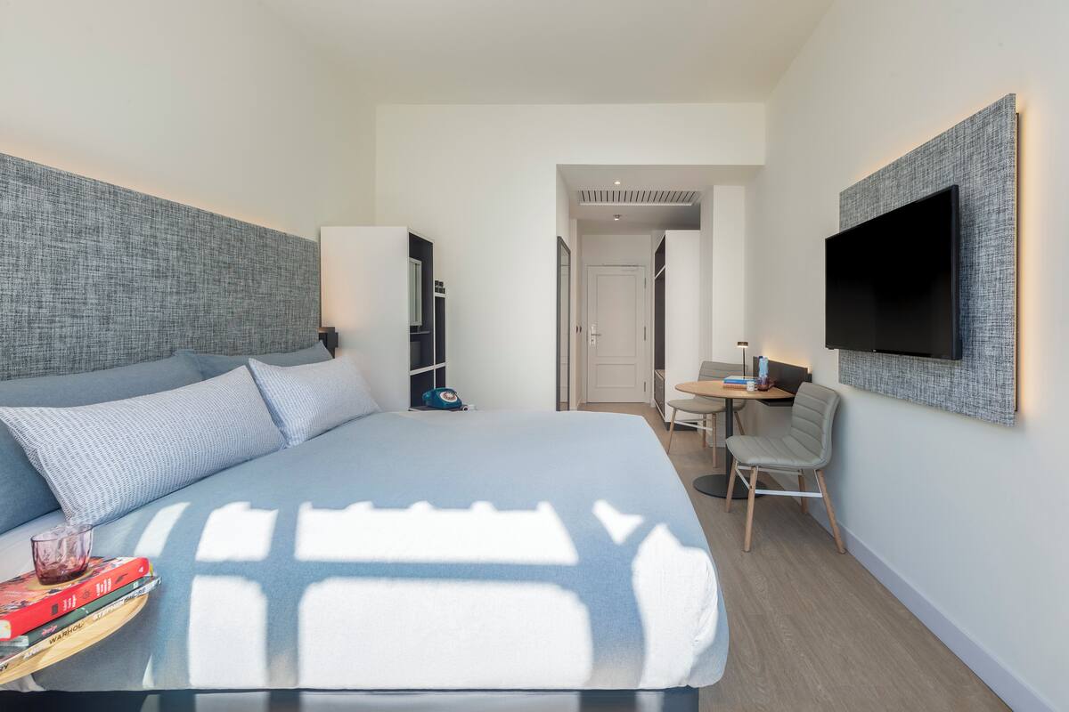 Ein modernes Hotelzimmer mit einem großen Bett, einem flachbildfernseher an der Wand, einem Schreibtisch mit Stuhl und Blick auf den Flur.