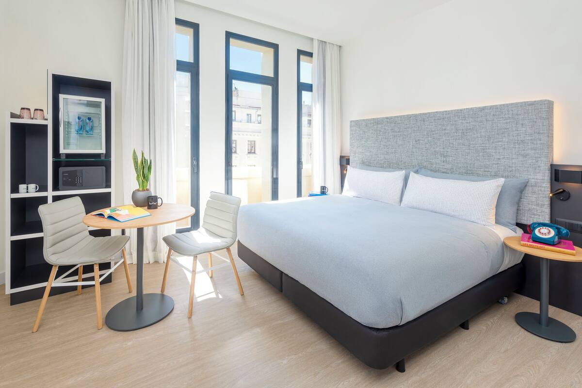 Ein modernes Hotelzimmer mit einem großen Bett, einem Schreibtisch mit Stuhl, einem kleinen Tisch und einem Bücherregal.
