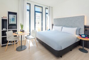 A modern hotel room with a large bed, a desk with a chair, a small table, and a bookshelf.