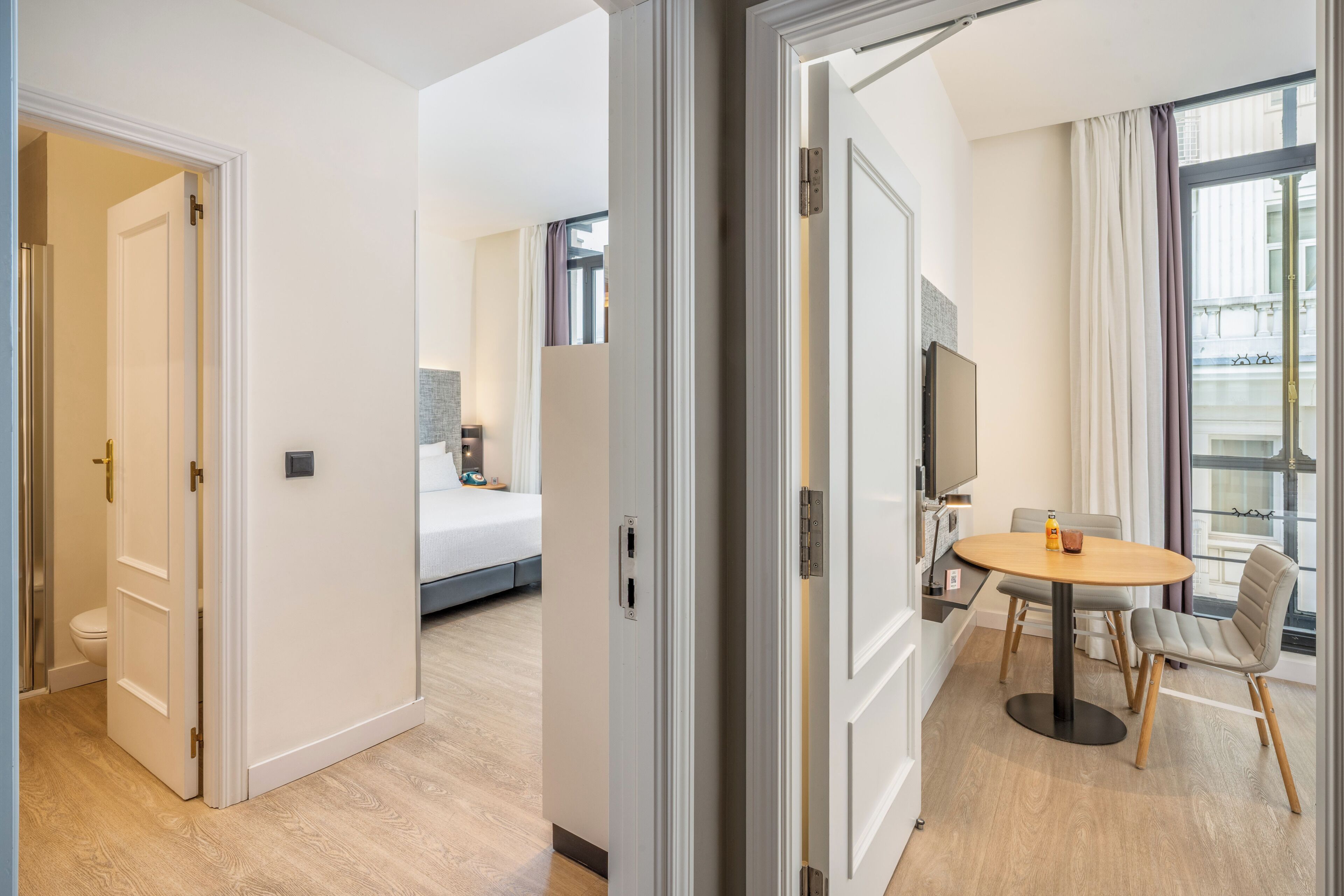 Habitación de hotel con cama, zona de comedor con mesa y sillas, televisión y ventana con cortinas.