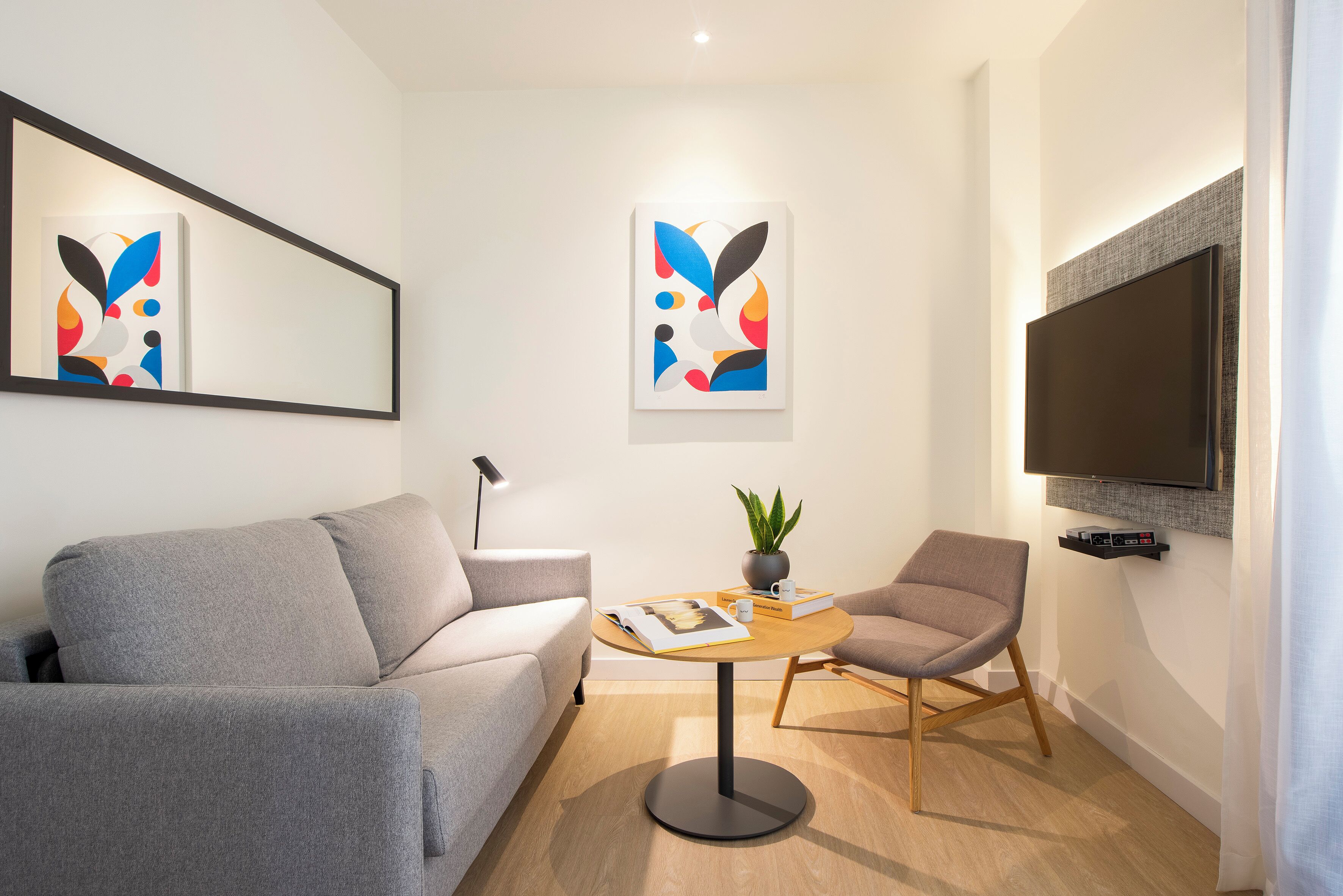 A modern living room with a grey sofa, a round wooden coffee table, and two armchairs. There are abstract artworks on the walls, a flat-screen TV mounted above the coffee table, and a potted plant on the table.