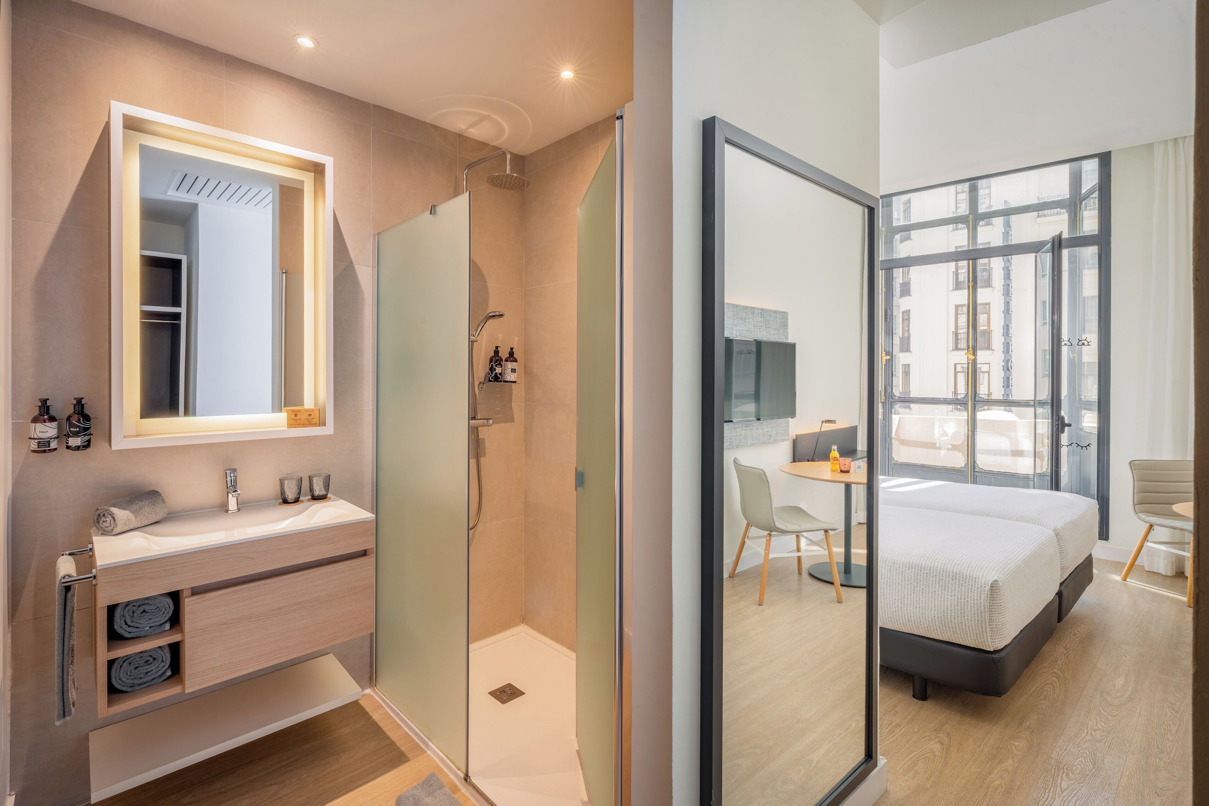 A modern bathroom with a glass shower enclosure, a vanity with a mirror, and a bedside table with a chair in the adjacent bedroom.