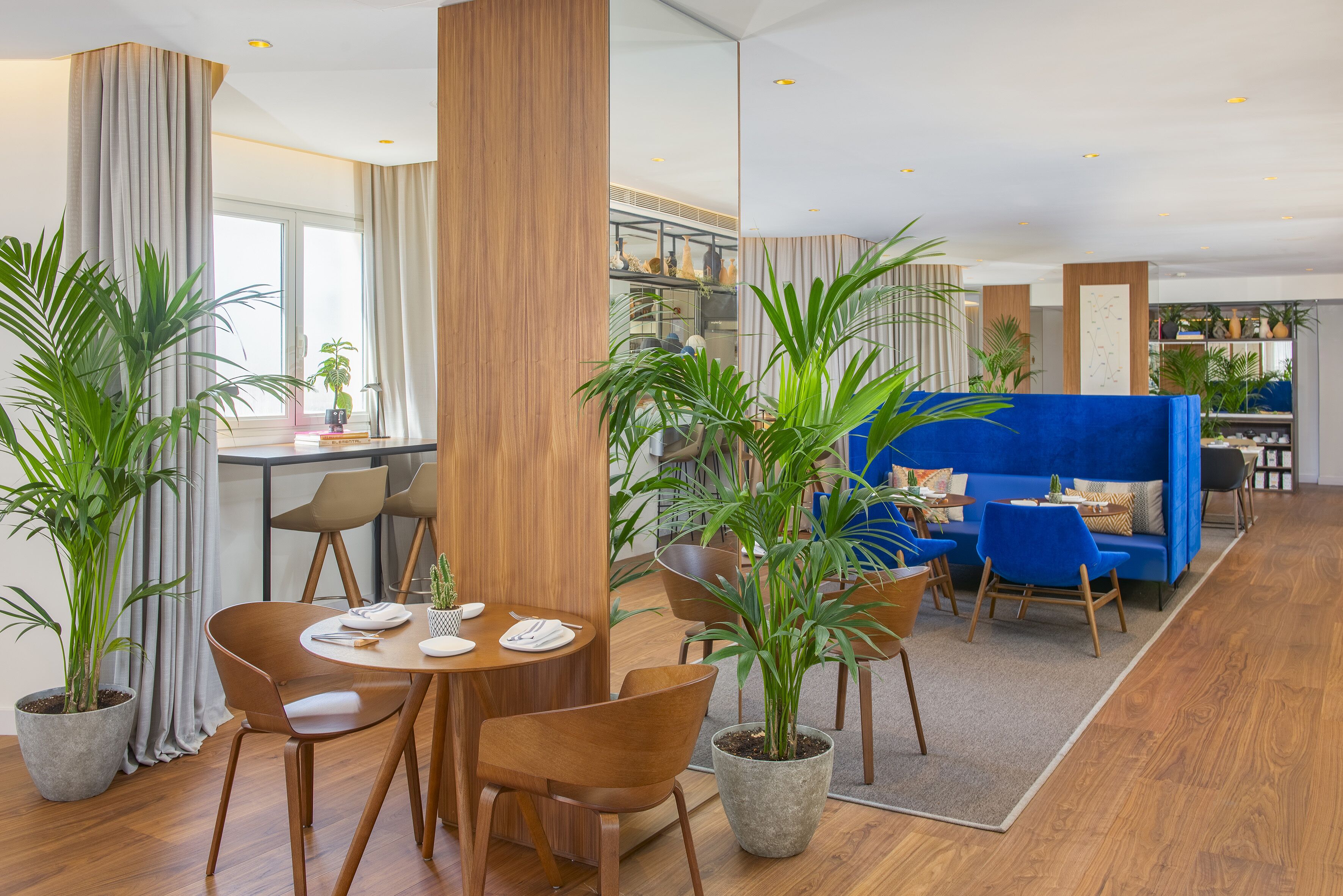 Un moderno hall de hotel con zona de comedor, sofá azul y plantas en macetas.