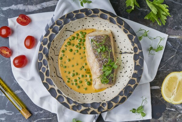 Un plato con filete de pescado, guisantes y salsa, acompañado de tomates cherry y perejil sobre una superficie de mármol.