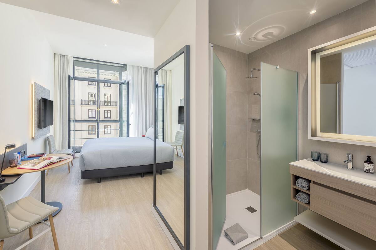 Una camera d'albergo moderna con un letto, una scrivania con una sedia, una TV, un'ampia finestra con tende, un box doccia e un angolo toilette con specchio.