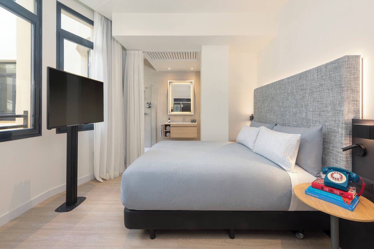 Una camera d'albergo moderna con un letto grande, una televisione, un comodino con libri, un bagno con uno specchio e una finestra con tende.