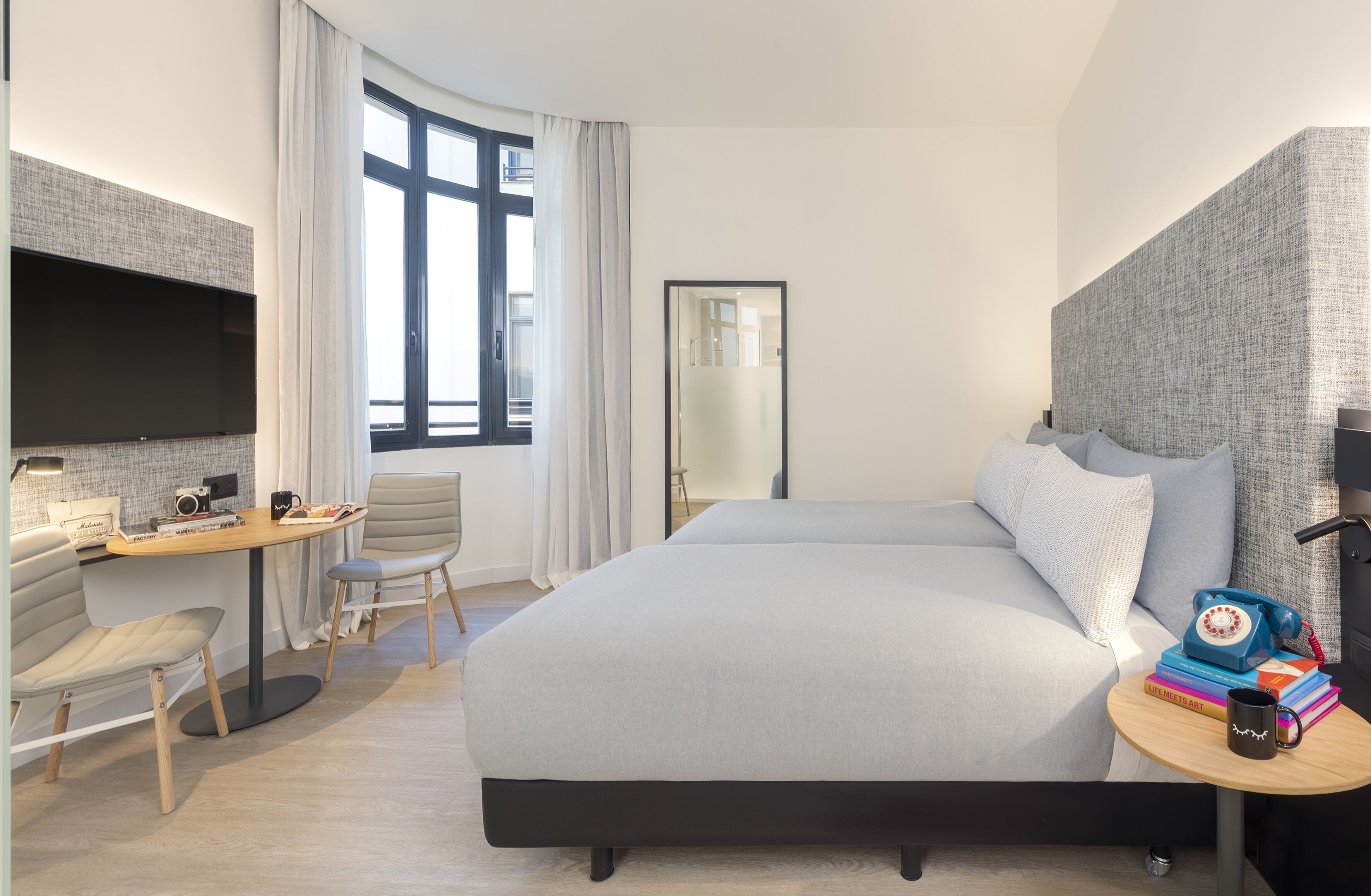 Moderni hotellihuone, jossa on suuri sänky, työpöytä tuolilla, taulutelevisio sekä pieni pöytä, jossa on puhelin ja kirjoja.