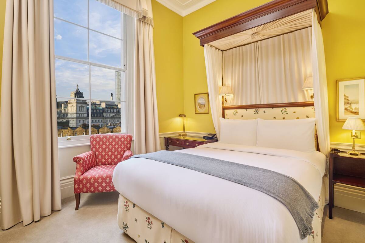 A hotel room with a large bed, a red patterned armchair, a nightstand with a lamp, and a view of a cityscape through a window with curtains.
