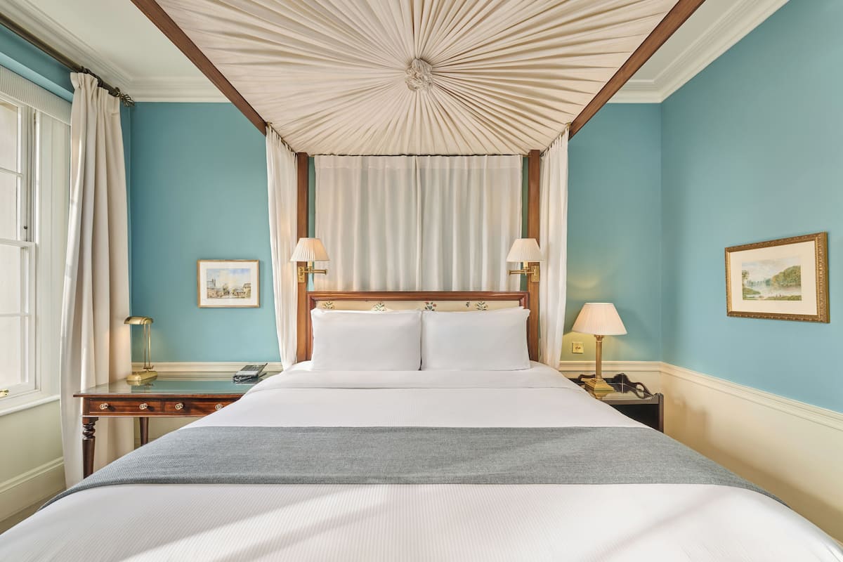 A neatly arranged bedroom with a large bed, a wooden headboard, two bedside lamps, a nightstand, and a window with curtains.