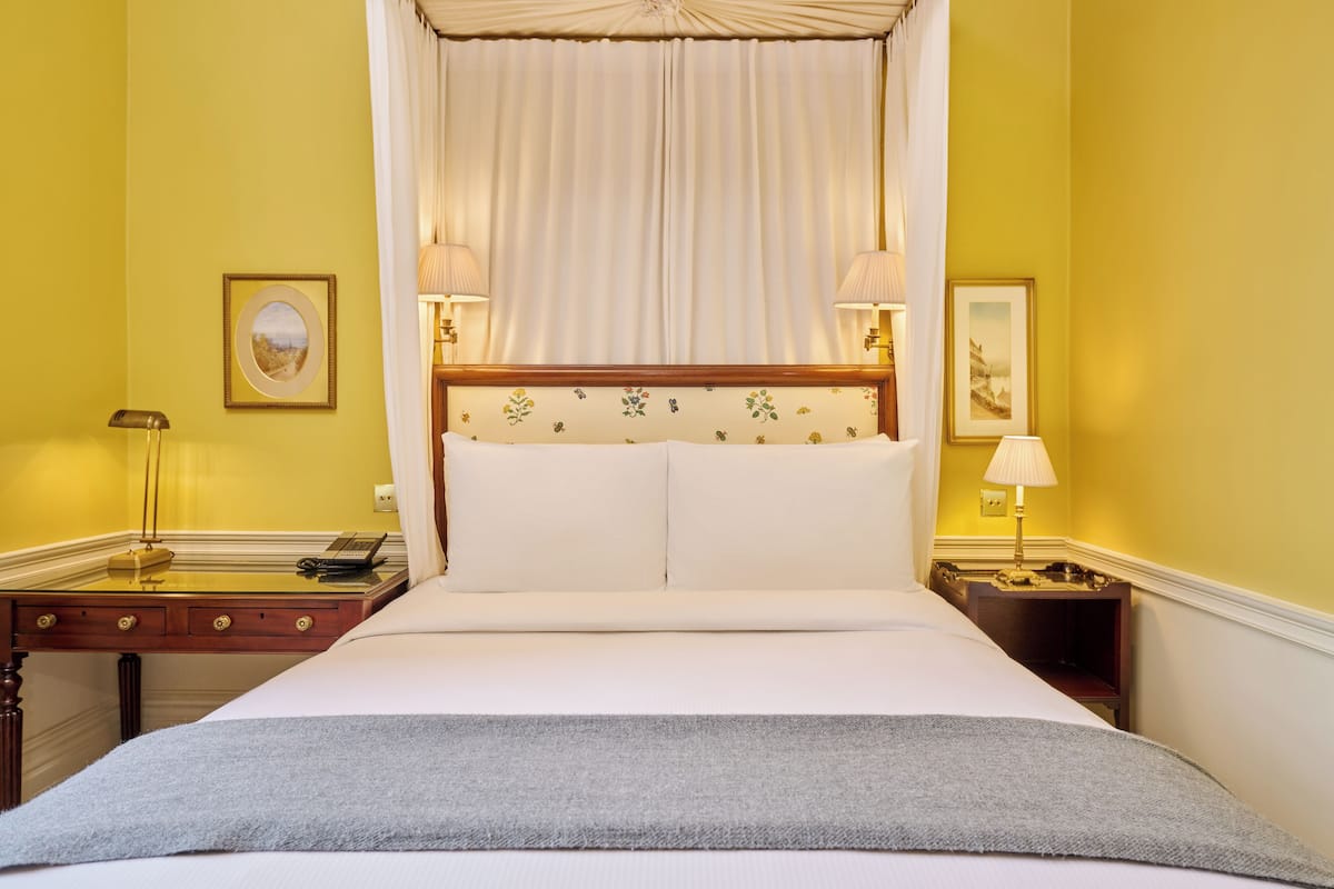 A hotel room with a large bed, a bedside table with a lamp, a desk with a lamp, and two framed pictures on the walls.