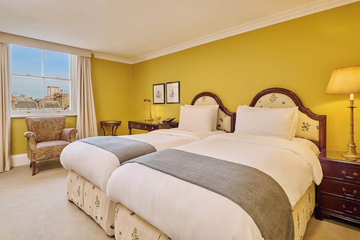 A hotel room with two beds, a chair, a nightstand, and a window with a view of buildings.