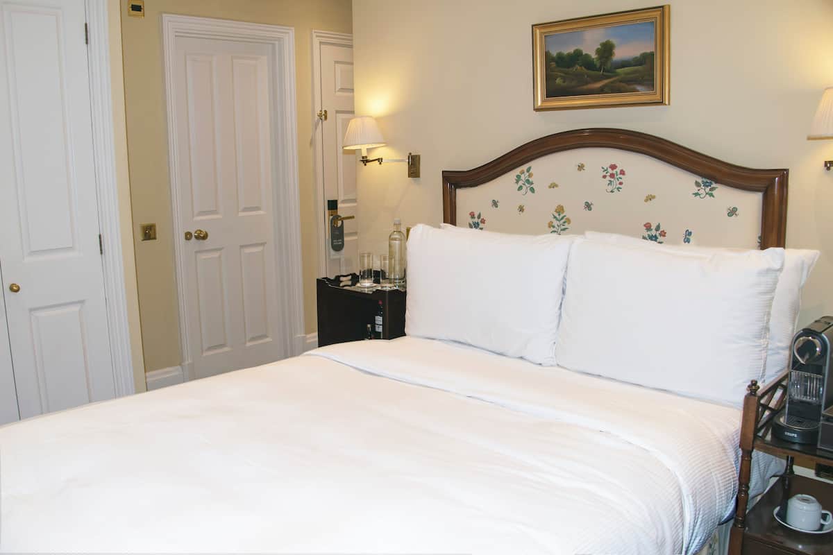 A neatly made bed with white linens, a wooden headboard with floral patterns, a bedside table with a lamp, and a painting on the wall.