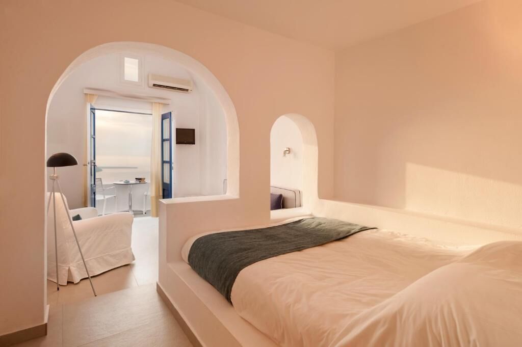 A bedroom with a bed, a desk, and a chair. There is a window with curtains and an air conditioning unit.