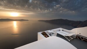 A white, modern outdoor seating area with a glass table and chairs overlooking a calm sea and distant mountains during sunset. - Regina Mare Adults Only (Santorini)