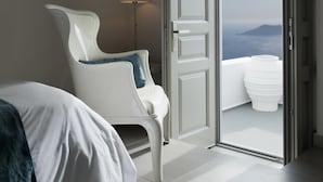 A hotel room with a bed, a white chair, a floor lamp, and a door leading to a balcony with a view. - Regina Mare Adults Only (Santorini)