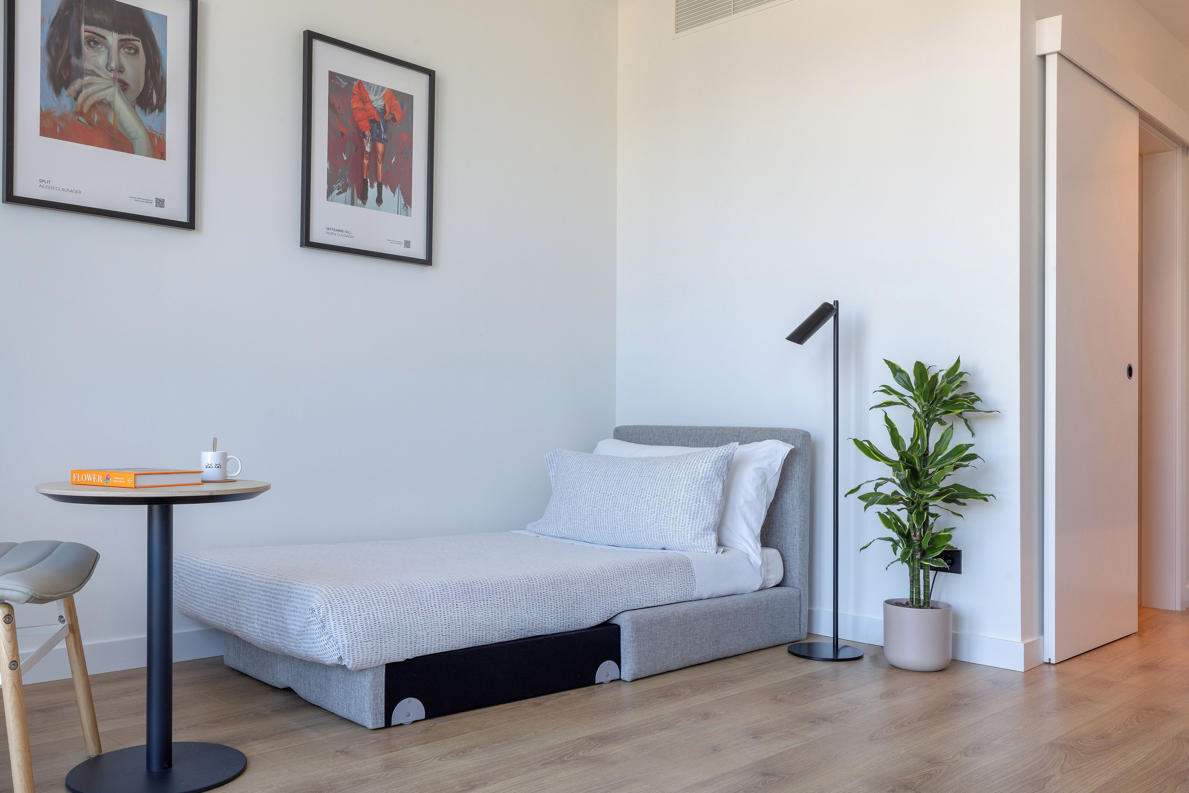 A minimalist room with a bed, a small table, a chair, a floor lamp, and a potted plant.
